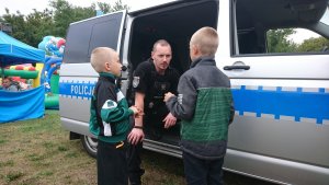 Na zdjęciu ukazany jest policjant siedzący w busie policyjnym, który to rozmawia z dwoma dziećmi. W tle widać także dmuchany zamek do zabaw dla dzieci.