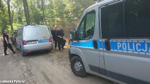 Na zdjęciu widoczne są dwa policyjne busy stojące w lesie. Obok nich policjanci, którzy przygotowują się do przeszukania lasu w celu odnalezienia zaginionego seniora.