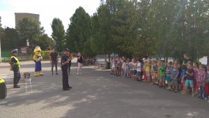 Na zdjęciu widoczny jest apel dla dzieci na boisku szkolnym. Dzieci stoją w szeregu, słuchają mówiącego do nich policjanta. W tle widzimy także człowieka maskotke.