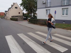 Na zdjęciu widzimy młodą kobietę, która przechodząc przez przejście dla pieszych, używa telefonu komórkowego, jednocześnie nie zwracając żadnej swojej uwagi na otoczenie.