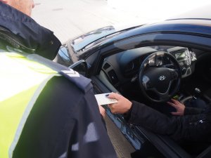 Zdjęcie przedstawia policjanta przyjmującego dowód rejestracyjny od kierowcy siedzącego w samochodzie.