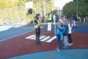 Zdjęcie przedstawia policjantkę rozmawiającą z dziećmi obok makiety przejścia dla pieszych na boisku szkolnym.
