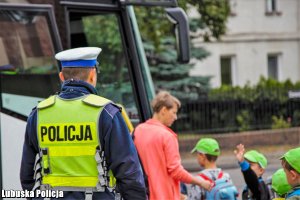 policjant stojący tyłem,  tle osoba dorosła z dzieckiem przy autobusie