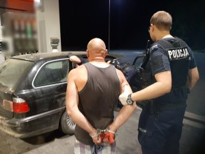 policjant trzymający osobę w kajdankach zapiętych z tyłu, w tle samochód osoby zatrzymanej
