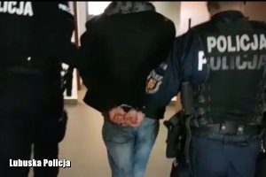 Dwóch policjantów prowadzi korytarzem zatrzymanego mężczyznę, który ma ręce skute kajdankami.