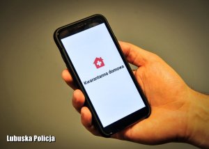 Dłoń trzymająca telefon na którym wyświetlony jest napis Kwarantanna Domowa oraz logo aplikacji