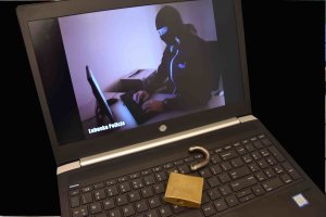 Laptop z pękniętą kłódką na klawiaturze, na monitorze wyświetlona postać osoby w kominiarce piszącej coś na klawiaturze