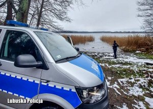 Policyjny radiowóz, a w tle policjant sprawdza czy nie ma żadnych osób na zamarzniętym jeziorze.