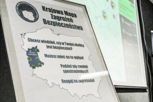 Zdjęcie przedstawia plakat krajowej mapy zagrożeń bezpieczeństwa na którym jest zarys granic polski z nazwą aplikacji a w tle wyświetlona mapa zagrożeń